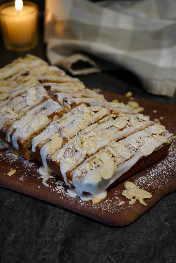 Sliced almond banana bread topped with white icing, powdered sugar, and slivered almonds on a wooden board, with a lit candle in the background.