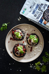 Overhead view of three blue corn sopes topped with refried beans, crumbled chorizo, queso fresco, and cilantro on a beige plate, with a bag of organic blue corn masa flour and scattered cheese around.
