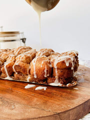 Close-up of Monkey Bread drizzled with vanilla icing as it’s being poured from above, sitting on a wooden board with visible cinnamon-sugar coating.