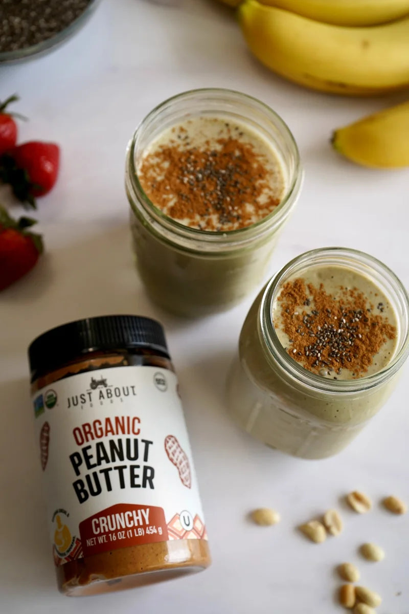Two green breakfast smoothies topped with cinnamon and chia seeds beside peanut butter and bananas