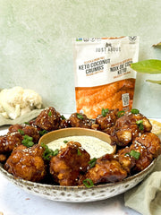 Bowl filled with teriyaki crispy cauliflower garnished with sesame seeds and green onions, served with a creamy dipping sauce, with a bag of keto coconut crumbs in the background